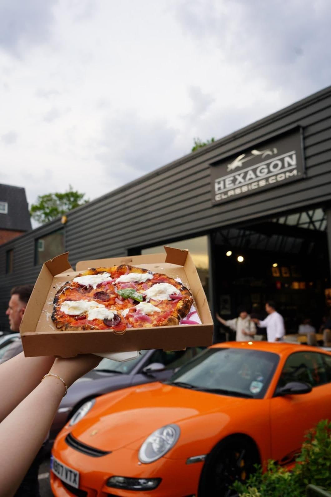 A boxed pizza held up beside an orange Porsche outside the showroom