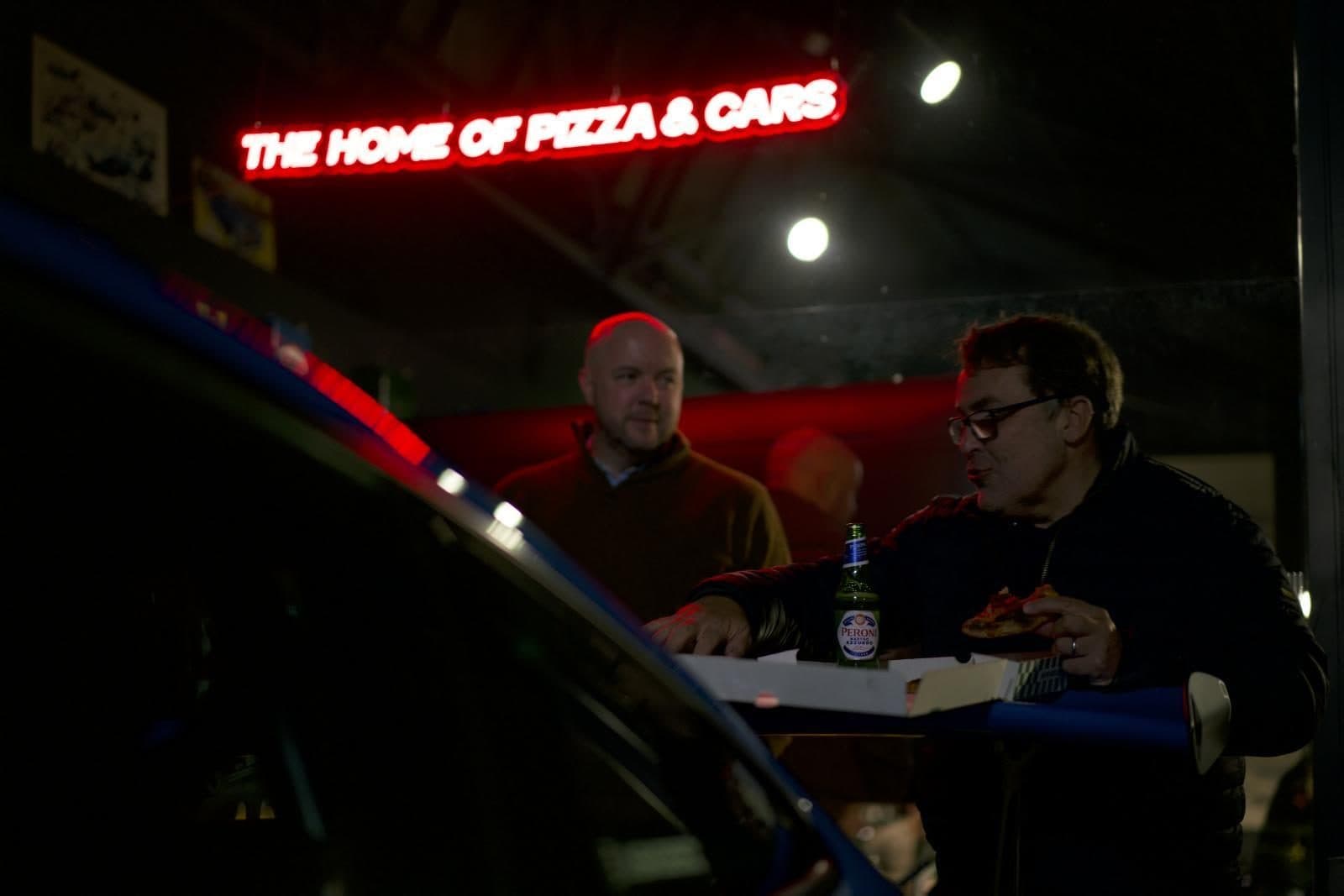 Guests eating pizza under the red neon 'The Home of Pizza & Cars' sign